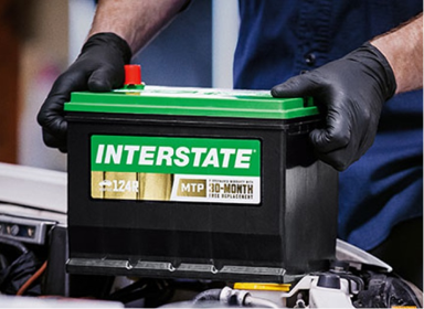 Technician wearing blue button-up and black rubber gloves grabbing onto a green and black Interstate car battery by the sides.