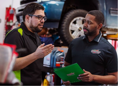 Two technicians are inside an auto shop. One of the men is speaking while raising his right hand to the other technician wearing an Interstate Battery uniform holding a green folder.