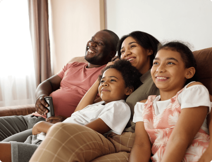 Interracial family of four sitting on brown sofa all smiling watching tv in a room with white walls and light brown curtains.