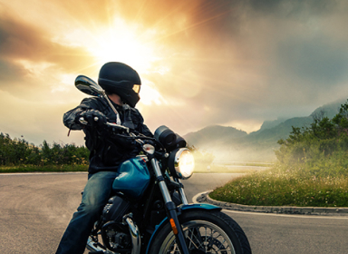 Person wearing black helmet, a black leather jacket, black gloves, and blue jeans is riding a blue motorcycle with the headlights on. The person riding the motorcycle has stopped his motorcycle and is looking to their left.
